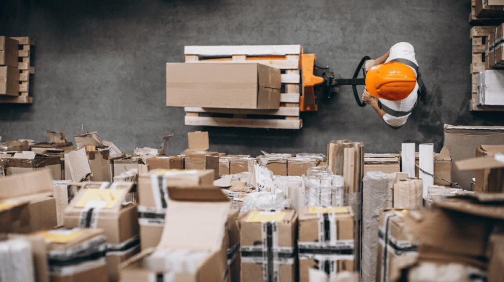 Overhead view of a warehouse worker in an orange hard hat maneuvering a pallet jack among stacked cardboard boxes
