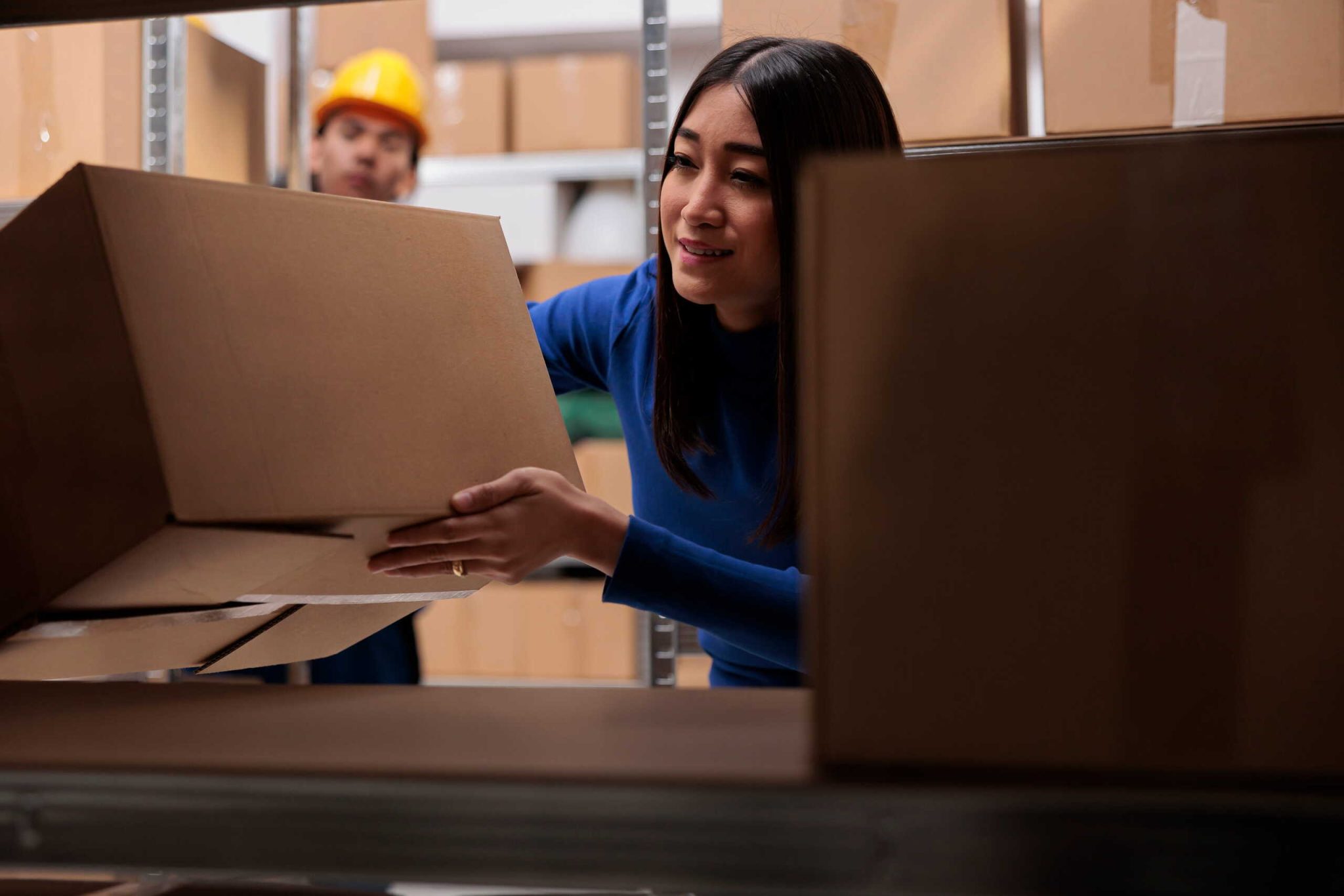 Female logistics worker placing a cardboard package onto storage shelving with a male worker in a hardhat in the background