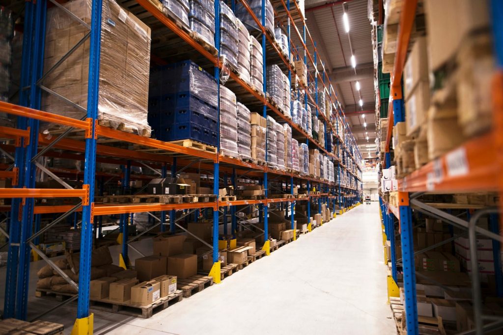 High-bay warehouse aisle lined with tall blue and orange heavy-duty pallet racking filled with inventory